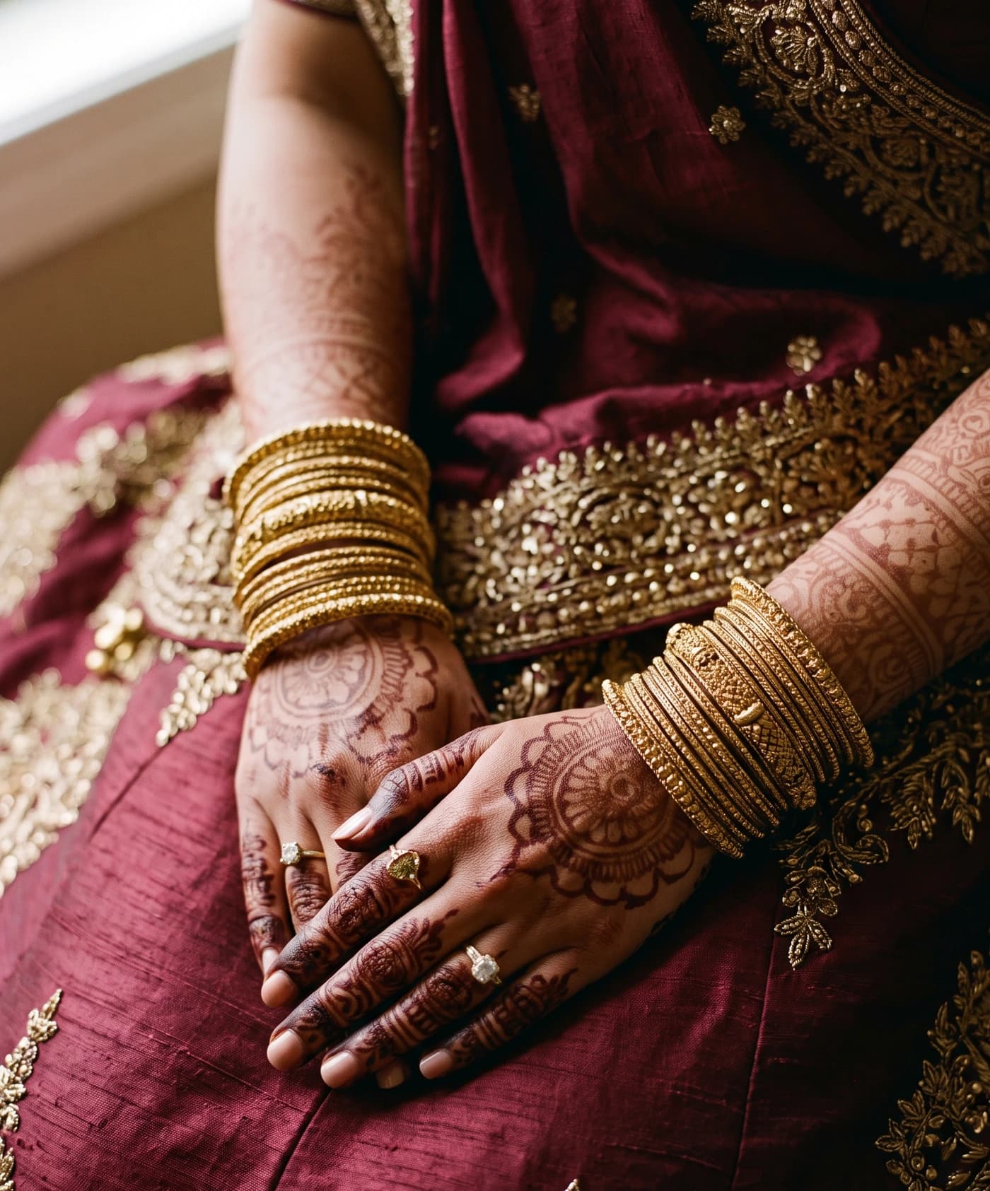 A bride's hands resting in her lap, patterned with mehendi and stacked with gold bangles, against a maroon and gold bridal outfit.